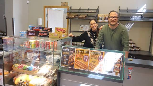 Två personer står bakom disken i en liten butik som säljer snacks, drycker och smörgåsar, med olika produkter utställda på hyllor och i glasmontrar.