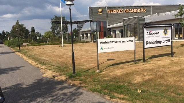 En byggnad med skyltar där det står ”Ambulansverksamheten” och ”Räddningsprofil” står på en gräsmatta nära en väg, med en flaggstång och en molnig himmel i bakgrunden.