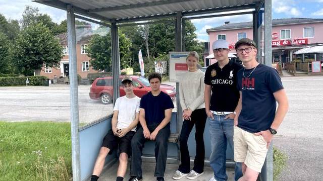 Fem unga människor står och sitter vid en busshållplats under tak en solig dag, med byggnader, bilar och träd i bakgrunden.