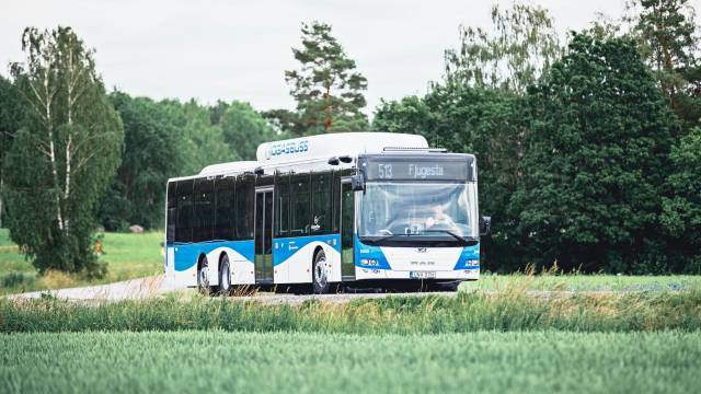 En blå och vit buss med beteckningen ”513 Fjugesta” kör på en landsväg som kantas av gröna åkrar och träd.