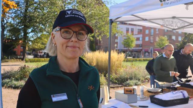 En kvinna med glasögon, namnskylt och keps står utomhus vid ett stånd med matbrickor. I bakgrunden syns andra personer nära ett vitt tält.