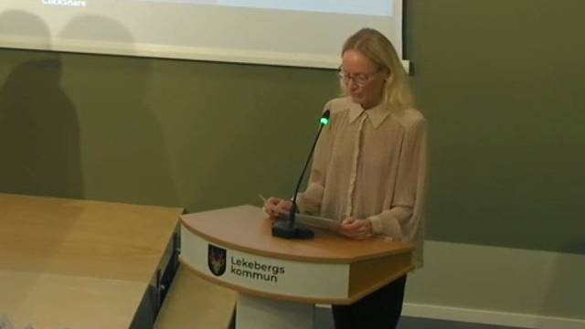 En person står vid ett talarstativ med texten ”Lekebergs kommun” och talar i en mikrofon i ett konferensrum.