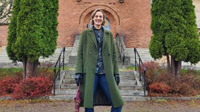 En person i grön kappa står på en stentrappa framför en tegelbyggnad, med ett stängt paraply och flankerad av två höga träd.