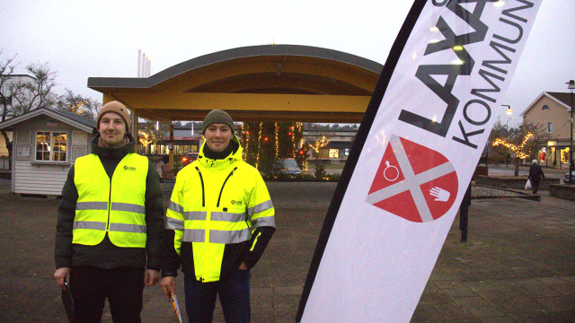 Två personer i gula reflexvästar står bredvid en flagga med texten 
