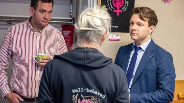 Tre personer samtalar inomhus; en man håller i en mugg, en annan är klädd i kostym och en person som står med ryggen mot kameran bär en huvtröja med en slogan.