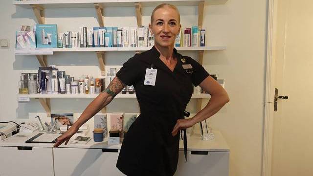 En kvinna i svart uniform poserar med ena armen utsträckt på en hudvårdsklinik, stående framför hyllor fyllda med skönhets- och hudvårdsprodukter.