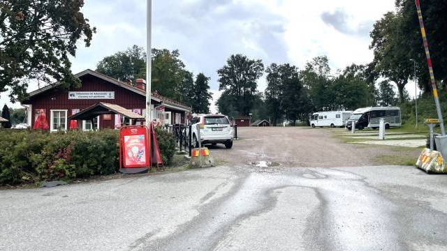 En bil närmar sig en liten röd byggnad med en skylt, omgiven av träd och parkerade husvagnar under en molnig himmel.
