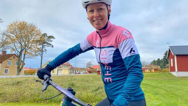 En person i cykelkläder och hjälm ler mot kameran medan han håller i en cykel utomhus i ett bostadsområde.