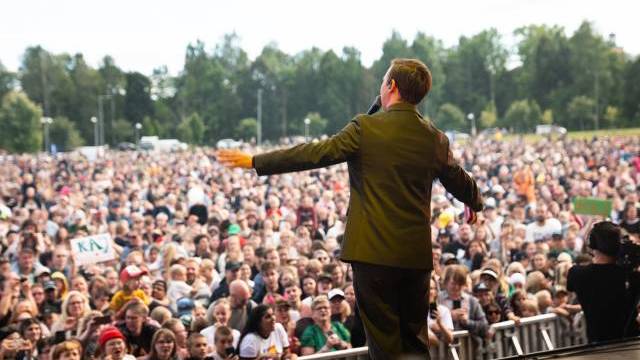 En person i kostym talar eller uppträder på en utomhusscen inför en stor publik, med träd och himmel i bakgrunden.