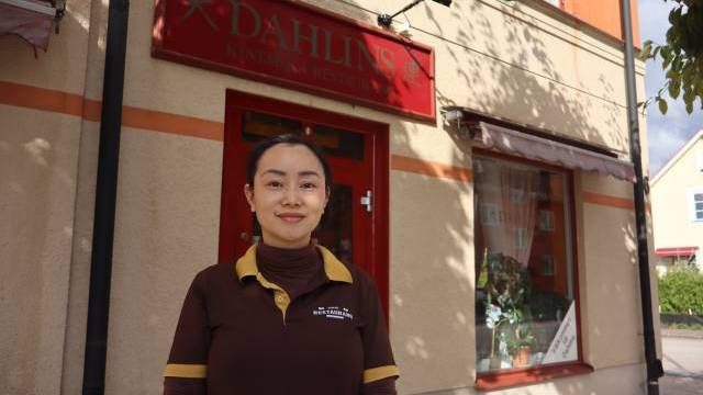En kvinna i brun uniform står framför en beige byggnad med en röd skylt där det står ”Dahlins Kinesiska Restaurang”.