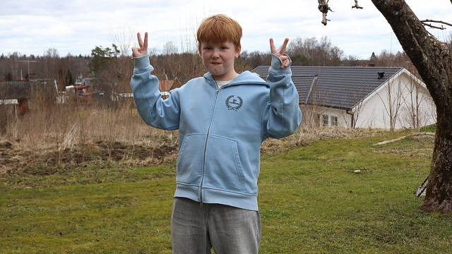 Ett barn med rött hår och ljusblå huvtröja står utomhus på en gräsmatta och håller upp båda händerna i fredstecken. Träd och hus syns i bakgrunden.
