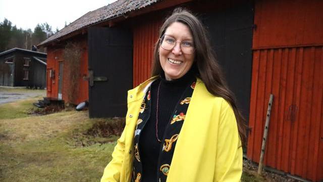 En kvinna i gul regnrock och glasögon står utanför en röd träbyggnad en molnig dag.