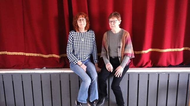 Två kvinnor sitter sida vid sida på en scenavsats framför fördragna röda gardiner.