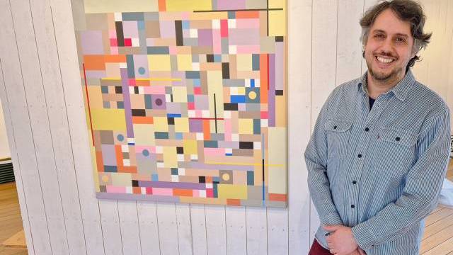 En man står och ler bredvid en färgglad geometrisk abstrakt målning på en vit trävägg.