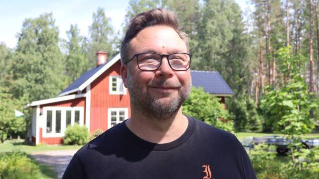 En man med glasögon och skägg står utomhus framför ett rött trähus omgivet av träd och grönska en solig dag.