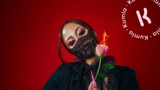 En kvinna i svart spetsklädsel och ansiktsskydd håller en brinnande rosa ros mot en röd bakgrund, med ordet 