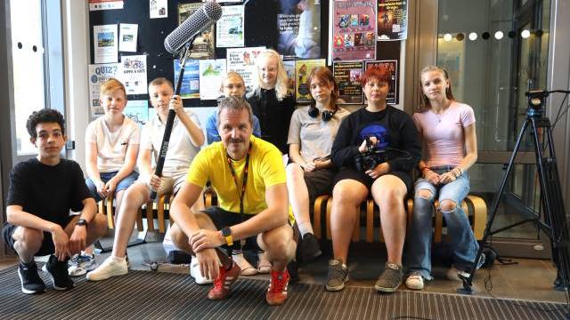 En grupp bestående av åtta ungdomar och en vuxen man poserar inomhus med filmutrustning framför en anslagstavla täckt av affischer.