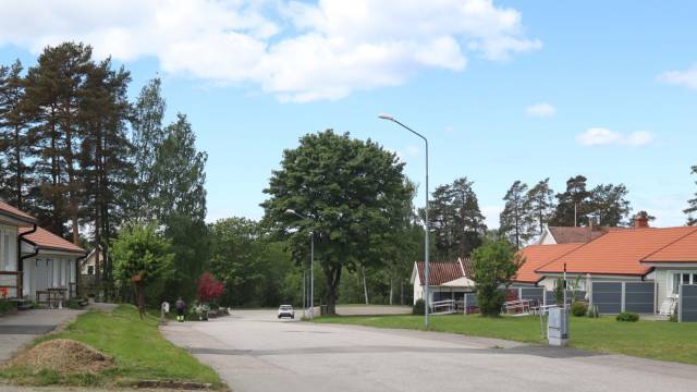 En lugn gata i en förort med hus, träd, en trottoar och en ensam bil som kör under en delvis molnig himmel.