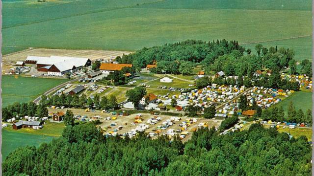 Flygbild över en stor campingplats med många tält och fordon, omgiven av träd och åkrar, samt flera byggnader på området.