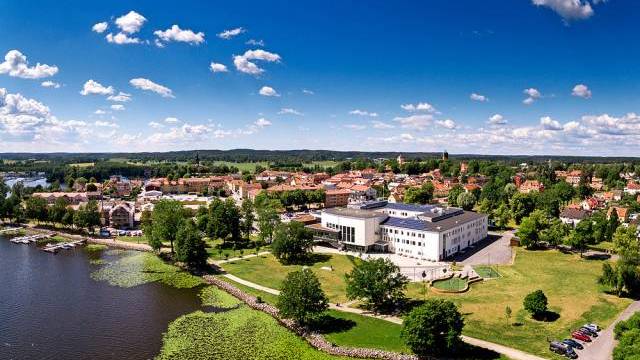 Flygfoto över en modern vit byggnad nära en flod, omgiven av gröna gräsmattor, träd och en stadsbild under en blå himmel med spridda moln.
