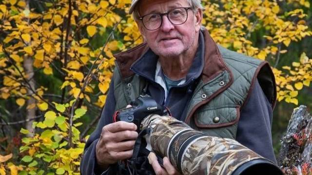 En äldre man med glasögon och friluftskläder håller i en Canon-kamera med ett stort kamouflageobjektiv och står framför höstlövverk.