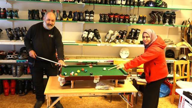 Två personer spelar på ett litet biljardbord i ett rum med hyllor som rymmer sportutrustning, inklusive hjälmar, skridskor och skyddsutrustning.