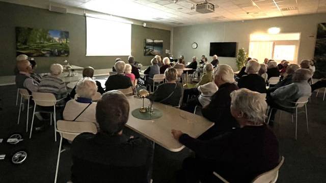 En grupp äldre personer sitter vid bord i ett svagt upplyst rum och tittar på en presentation på en projektorskärm längst fram.