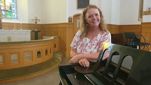 En kvinna i en blommig blus står bredvid ett piano i en kyrka med träpaneler, ett målat glasfönster och ett altare i bakgrunden.