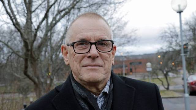 En äldre man med glasögon och mörk rock står utomhus i en parkliknande miljö med träd och en byggnad i bakgrunden.