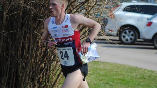 En manlig löpare med startnummer 24 och en karta i handen tävlar utomhus i närheten av buskar och parkerade bilar.