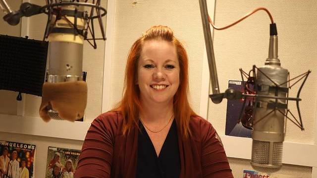 En kvinna med rött hår sitter vid ett skrivbord i en inspelningsstudio och ler mot kameran, med två stora mikrofoner framför sig.