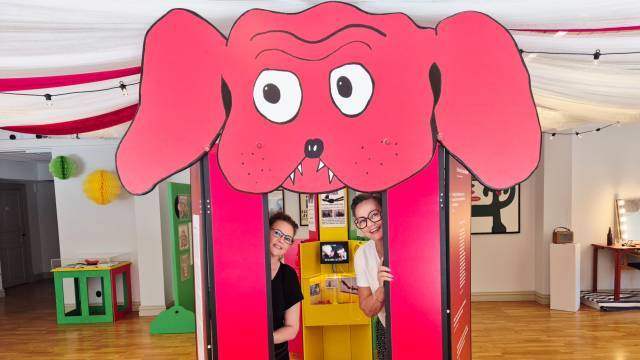 Två personer står inuti en röd utställningsfigur föreställande en tecknad hund med stora ögon och öron i ett starkt upplyst rum med trägolv och färgglada dekorationer.
