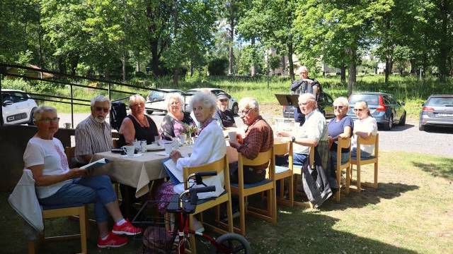 En grupp äldre sitter vid ett bord utomhus och njuter av en gemensam måltid. En person i bakgrunden spelar dragspel. Bakom dem syns träd och parkerade bilar.