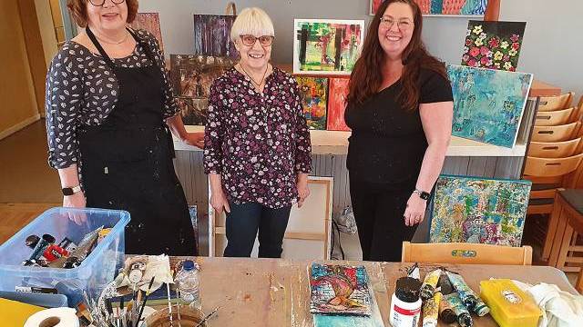 Tre kvinnor står och ler framför en utställning av färgglada målningar i en konststudio, med konsttillbehör och dukar på ett bord i förgrunden.