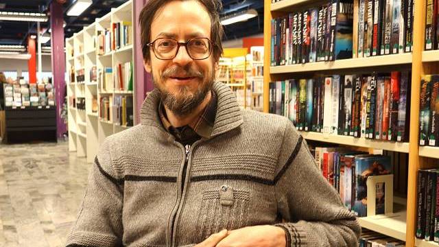 En man med glasögon och grå tröja sitter i en bokhandel och ler mot kameran, med bokhyllor i bakgrunden.