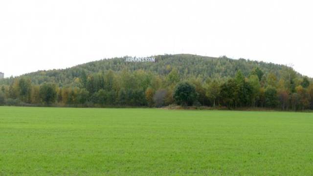 Ett brett, grönt fält som leder upp till en trädklädd kulle med några antenner på toppen under en molnig himmel.