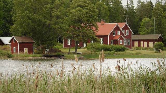 En grupp röda trähus med vita kanter ligger vid en liten sjö, omgiven av höga träd och vass i förgrunden.