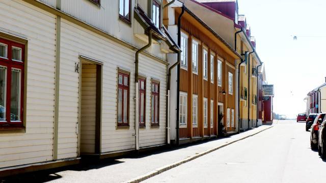 En lugn gata kantad av färgglada trähus i solskenet, med parkerade bilar på höger sida och en klar himmel ovanför.