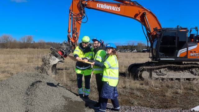 Tre personer i skyddsutrustning håller i spadar på en byggarbetsplats nära en grävmaskin som flyttar en grushög under en klar dag.
