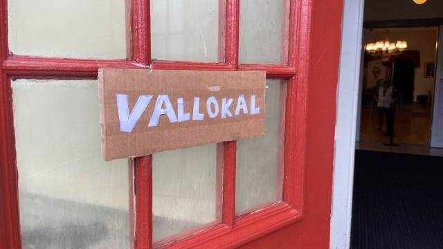 En handskriven skylt med texten ”Vallokal” är uppsatt med tejp på en röd dörr, vilket anger att det är en vallokal. Dörren står öppen och man kan se in i lokalen där det finns ljuskronor.