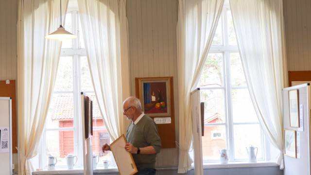 En äldre man står inomhus nära höga fönster med vita gardiner och håller i en inramad tavla. På fönsterbrädan syns en tavla och flera kannor.