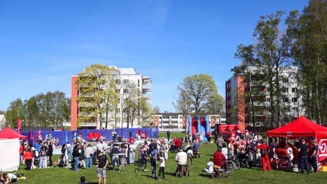 En folkmassa samlas på en gräsyta mellan hyreshus, med röda tält, ballonger och dekorationer under en klarblå himmel.