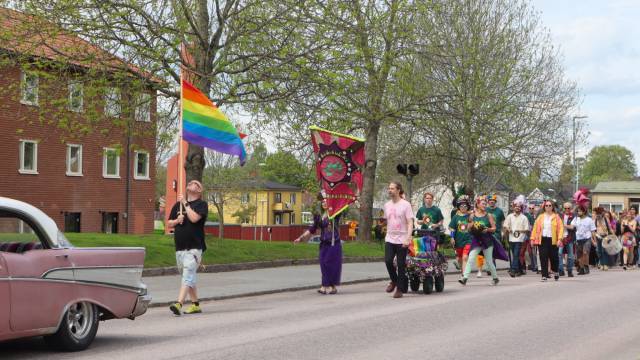 En grupp människor marscherar längs en gata i en parad, där en person håller i en regnbågsflagga och andra bär färgglada banderoller och har festliga dräkter.
