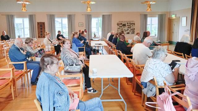 En grupp vuxna sitter på trästolar och vid bord i ett samlingsrum, vända mot framsidan där någon verkar hålla ett tal eller göra en presentation.
