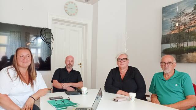 Fyra vuxna sitter vid ett vitt bord på ett kontor, med böcker, koppar och elektroniska apparater framför sig. I bakgrunden syns en väggklocka, en dörr och en landskapsmålning.