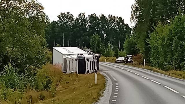 En stor vit lastbil ligger på sidan vid sidan av en kurvig landsväg, med träd och parkerade fordon i närheten.