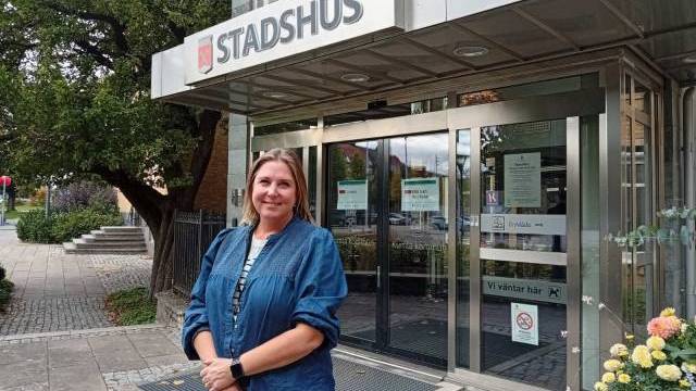 En kvinna står framför ingången till en byggnad där det står ”STADSHUS” på en skylt. Det finns glasdörrar, anslag och några blommor och träd i närheten.
