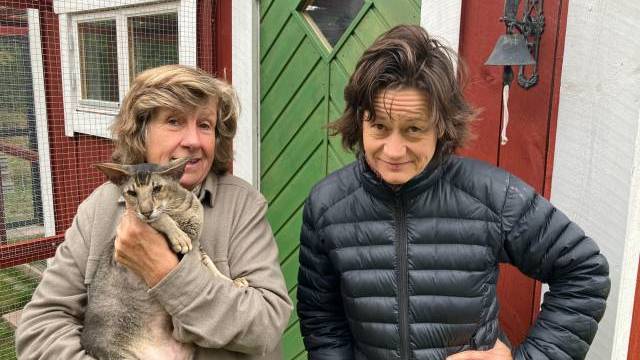 Två personer står utanför en röd och vit byggnad; den ena håller en katt, och den andra står med en hand på höften. I bakgrunden syns en grön dörr och en metallklocka.