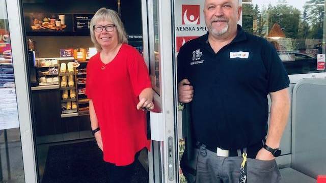 Två personer står vid ingången till en närbutik, en kvinna håller dörren öppen och en man står bredvid henne, båda ler mot kameran.