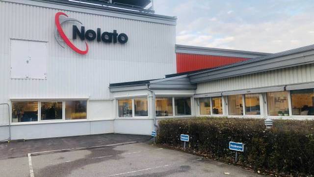 Exteriör av en Nolato-byggnad med stor logotyp, flera fönster och två reserverade parkeringsplatser på framsidan under en molnig himmel.